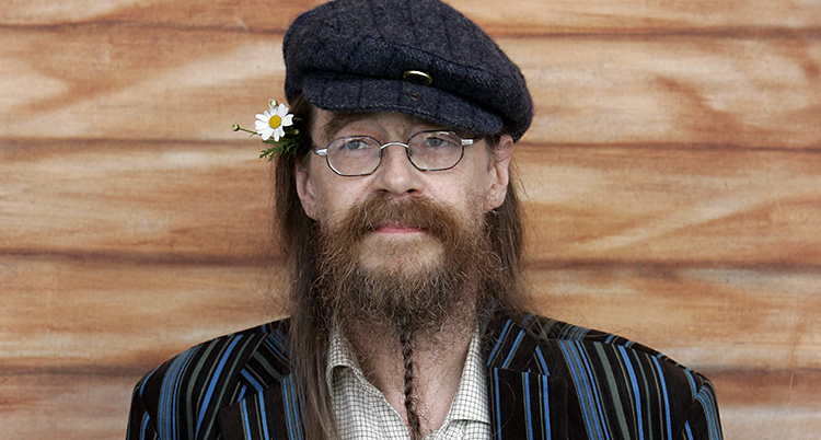 Han står vid en vägg. Han har långt hår, skägg och glasögon. Han har en keps på huvudet och en blomma bakom örat.
