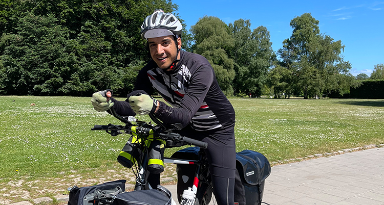 Han har svart cykelhjälm, svarta kläder och håller i styret på en cykel med händer i handskar. Han står utomhus. Det är grönt omkring honom.