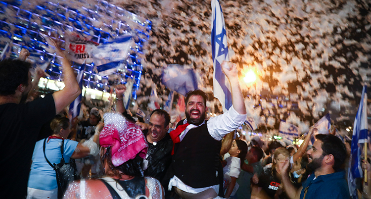 Det är kväll. Folk jublar. En man i väst och vit skjorta skrattar och håller upp en knuten hand. Bakom honom Israels flagga.