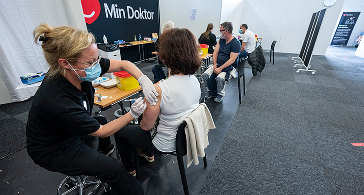 En kvinna med svarta kläder och munskydf gefr en spruta till en kvinna som sitter med ryggen mot kameran. Bakom dem sitter flera och ska få sprutor. d