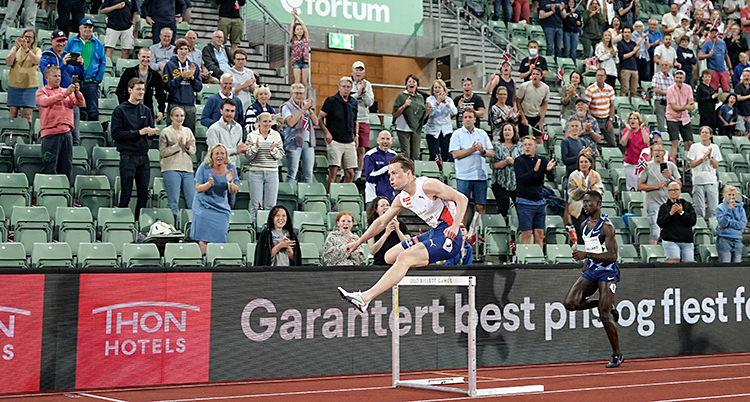 Från en stadion för friidrott. Karsten Warholm hoppar över en häck. På läktaren står publiken upp och hejar på honom.