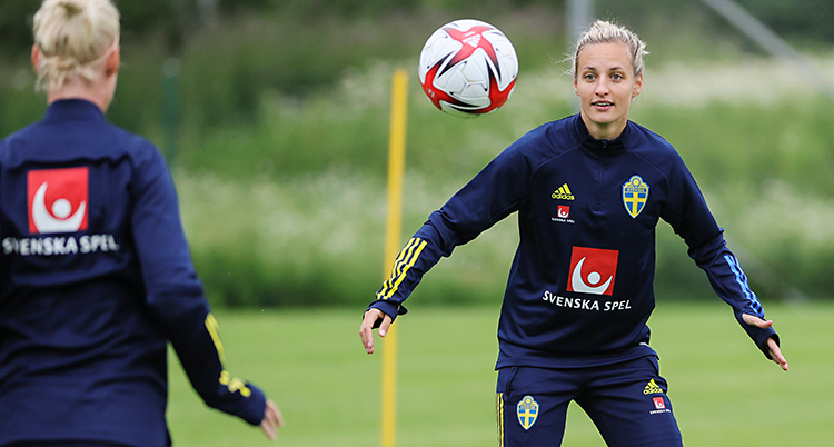 En träning för fotboll. Bollen är i luften. Nathalie Björn tittar på bollen och gör sig redo. Hon har blå kläder med den svenska flaggan på.
