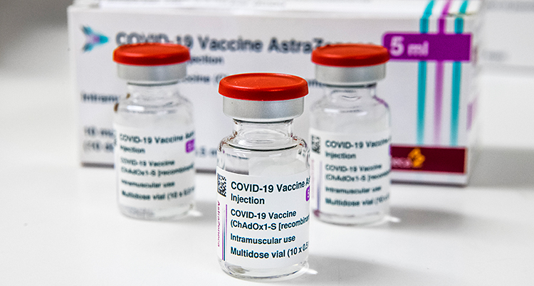 Tre små flaskor av glas. Det är vaccin i flaskorna. Flaskorna har lock som är röda. I bakgrunden står en liten papplåda.