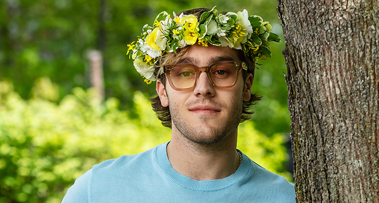 Han står vid ett träd. Han tittar in i kameran. Han har glasögon och en krans med blommor på huvudet.