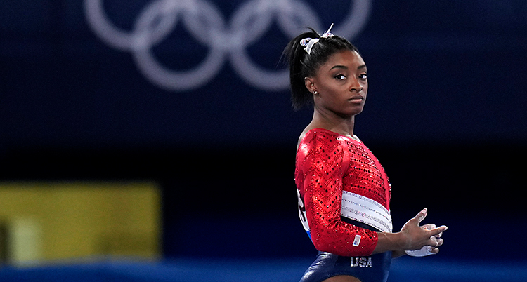 Simone Biles från en tävling i OS. Hon ser allvarlig ut. Hon har en dräkt som är röd, vit och blå.