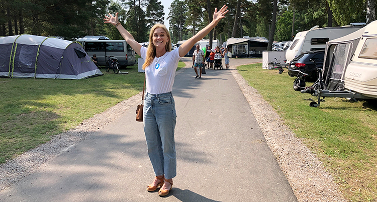 Hon står utomhus på en camping, omgiven av husvagnar. Hon sträcker armarna mot luften.