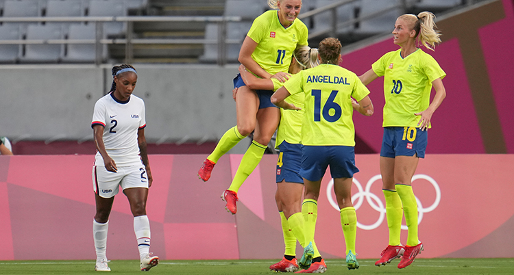 Hon har gul fotbollströja och hoppar upp i luften. Med kameran mot ryggen står en till spelare med gul tröja och en tredje i profil. De är glada. Till vänster går en spelare i vit tröja med besviken min.