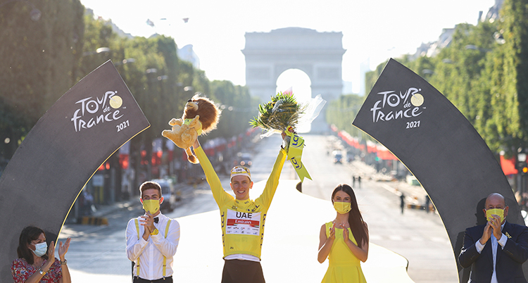 I mitten står en man med gul cykeltröja. Han sträcker armarna mot luften och håller i en pokal och en blombukett. Han flankeras av flera personer. De står utomhus, på en stor karta i Paris. Triumfbågen syns i bakgrunden.