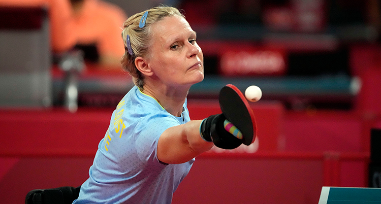 Hon har ljusblå tröja, ljust hår, och är på väg att slå en vit bordtennisboll med racket hon håller i höger hand.