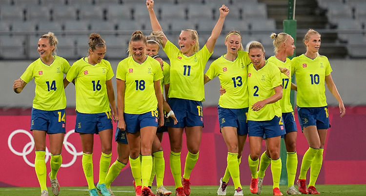De går på en rad, de har gula fotbollströjor, blå shorts och gula fotbollsstrumpor. Hon i mitten sträcker upp armarna i luften. De är på en arena.