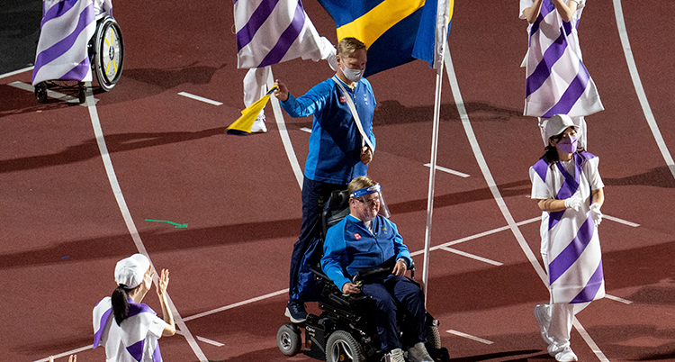 Han är i mitten av bilden, sitter i en rullstol och bär den svenska flaggan. Han har blå tröja. Han är på en arena.
