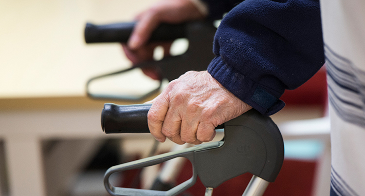 Två gamla händer håller i handtagen till en rollator.