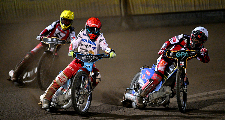 Tre speedwaymotorcyklar kör i en kurva på den grusiga banan. Förarna har dräkter och hjälmar med visir.