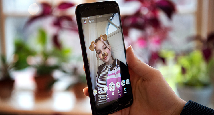 En hand håller i en mobil med en bild av en flicka. Bakom syns ett fönster med krukväxter.