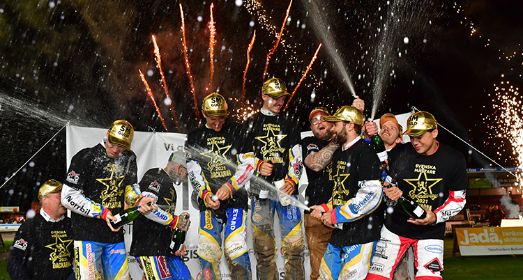 De står i en grupp på ett podium, de har guldhattar och öppnar champagne. Bakom syns fyrverkerier.