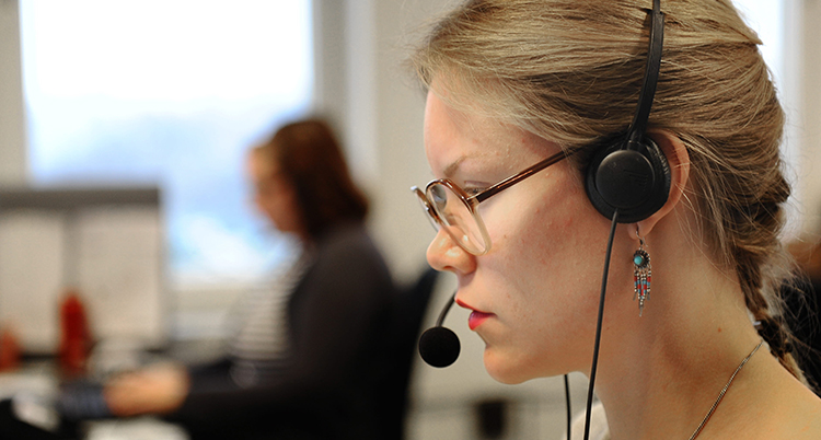 Hon har ett headset och syns från sidan. Bakom syns en person som sitter framför en dator och jobbar.