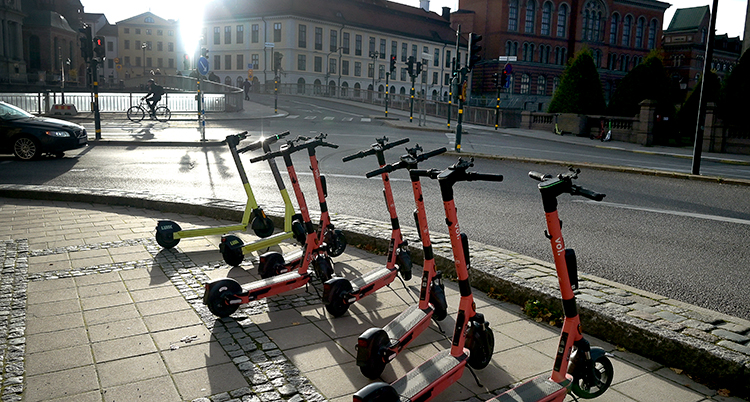Elsparkcyklar står parkerade intill varandra på en gata. Bakom syns en trafikkorsning och hus.