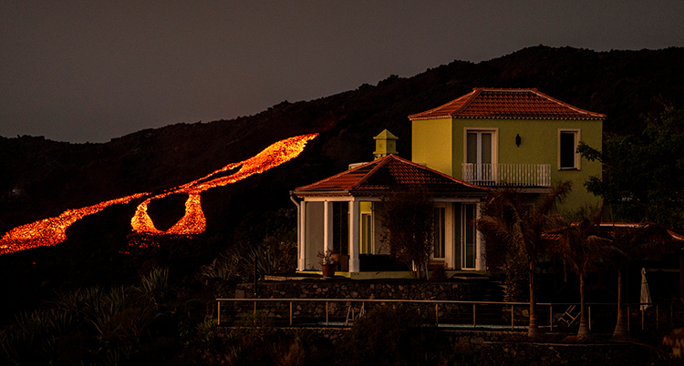 Ett hus i förgrunden, i bakgrunden röd lava som rinner nedför en bergssluttning. Det är mörkt ute.