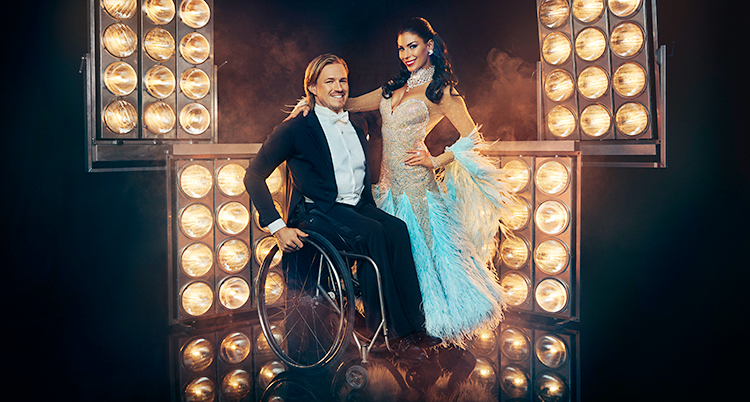 De har fina kläder. De ler och tittar in i kameran. Jasmine står upp. Aron sitter i rullstol.