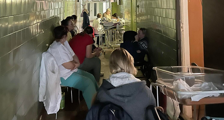 Kvinnor sitter i en korridor. Det står små sängar för barn i korridoren.