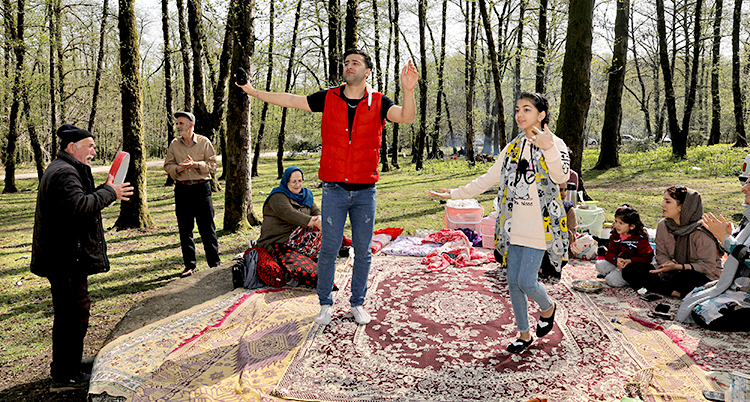 Människor har picknick i en skog. De har lagt ut mattor på marken. De umgås och dansar.