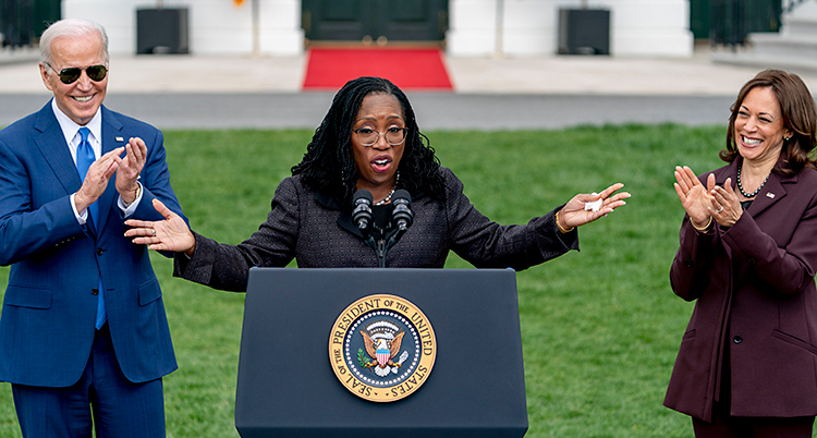 Hon står utomhus och håller ett tal i en talarstol. Till vänster om henne står Joe Biden. Till höger om henne står Kamala Harris.