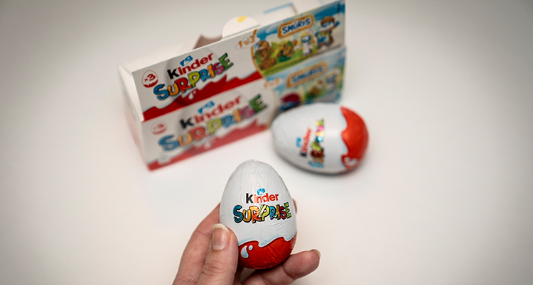 En hand håller ett oöppnat kinderägg. I bakgrunden ett till ägg och en ask för äggen.