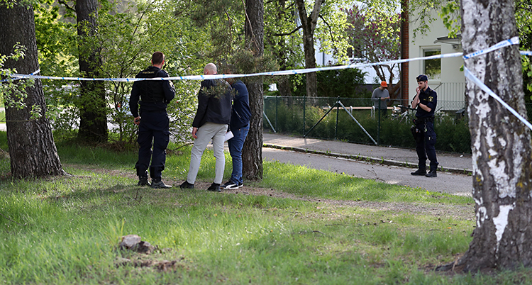 Bilden är tagen utomhus vid en liten park nära några hus med lägenheter. Några poliser står bakom avspärrningsband. Banden har satts upp mellan träd.