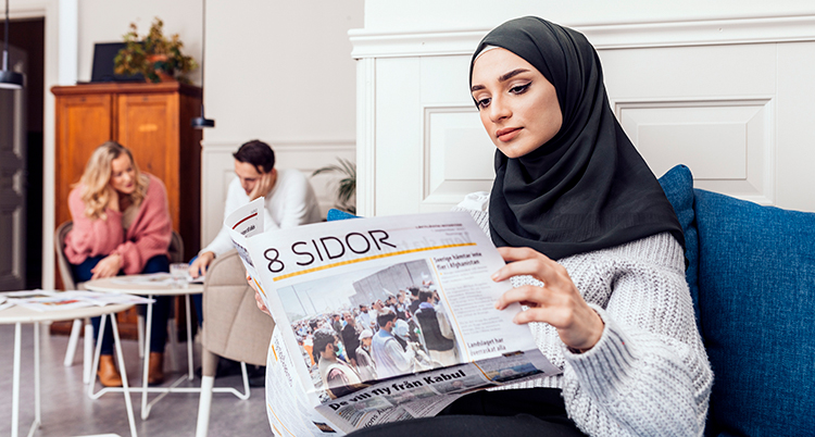 Hon sitter i en soffa och läser tidningen. Hon har svart slöja och en grå tröja. Bakom syns en kvinna och en man vid ett bord.