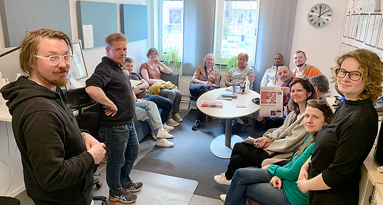 En grupp sitter runt ett bord och några står. Alla tittar in i kameran