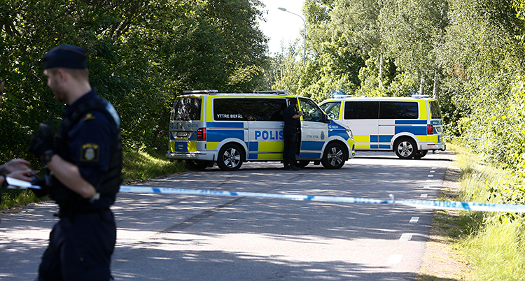 Poliser har spärrat av en väg. Flera polisbilar står på vägen. Det är soligt väder och grönska längs vägen.