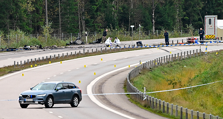 Några poliser jobbar på en vägg. Rester från en förstörd bil finns på vägen.