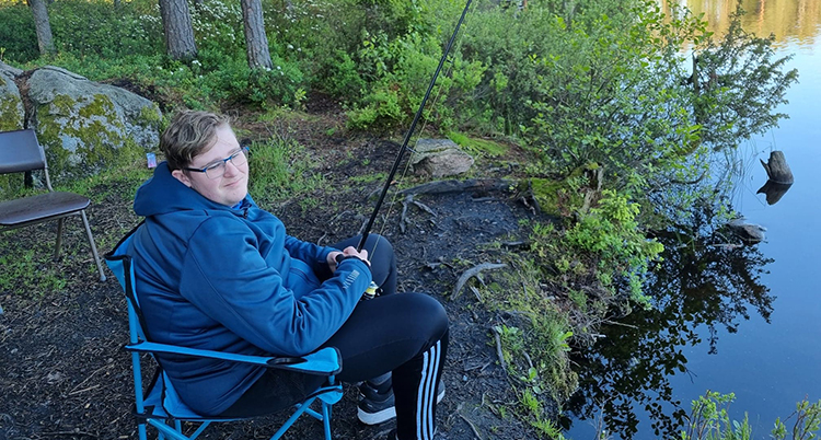 Han sitter i en stol intill en sjö, han håller i ett metspö. Han vrider huvudet mot kameran. Bakom syns gräs, vass och en klippa.