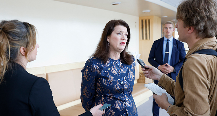 Hon står i mitten av bilden och pratar i en mikrofon som en man sträcker fram. Han står till höger. Till vänster står en kvinna. Bakom finns en vakt. De är utanför riksdagen.