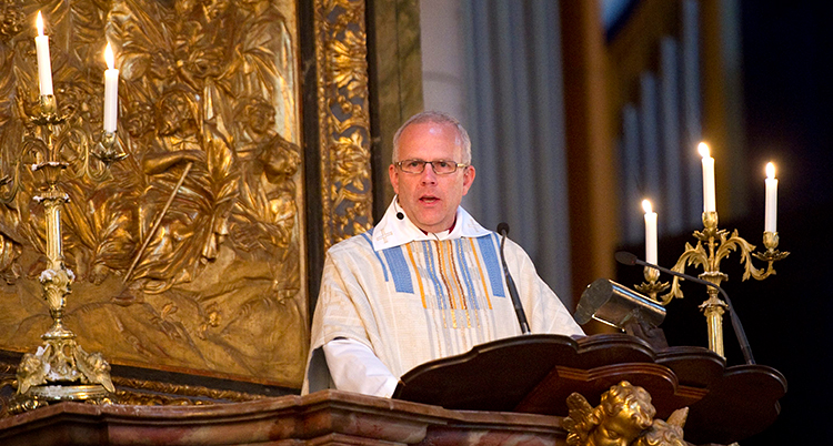 Han har en vit kåpa och står i en talarstol i en kyrka. Det brinner ljus i två ljusstakar.