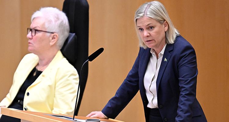 Hon står i en talarstol och pratar i en mikrofon. Bakom syns en träfärgad vägg. Till vänster syns en kvinna som sitter.