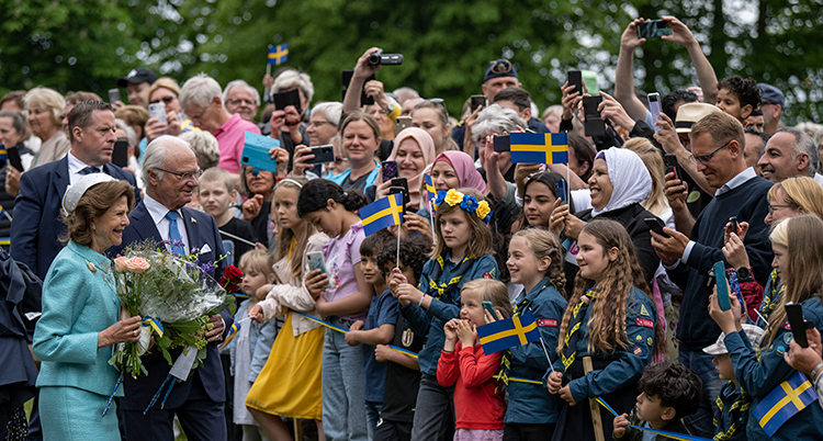 Massor av folk tittar på kungen och drottningen och viftar med flaggor