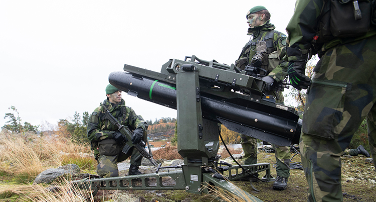Det är klippor och torrt gräs. En militär står bredvid en robot i ett ställ. En annan militär sitter på huk intill och tittar. Längst fram i bild syns ryggen på en tredje militär.