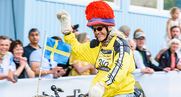Han har gul träningsjacka, en mössa med stor tofs, en flagga i cykelkorgen och han vinkar med handen som han har en vit vante på.