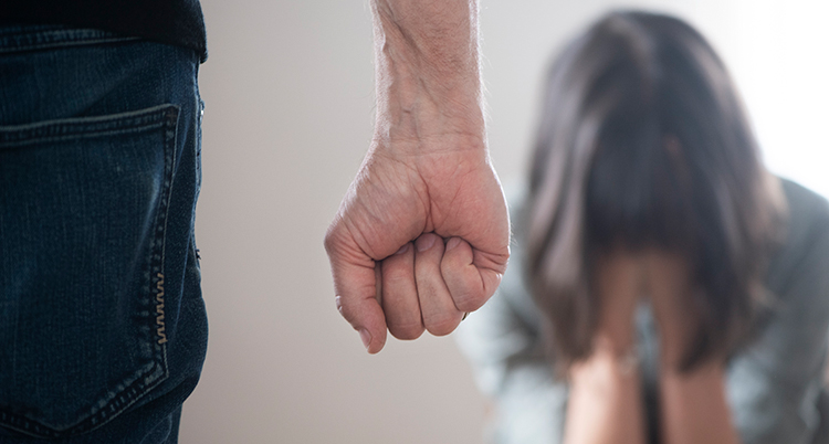 i förgrunden syns byxorna av en man med en knuten näve hängande längs sidan av benet. I bakgrunden syns en sittande person med händerna för ansiktet. Personen ser ut att gråta eller vara mycket rädd.