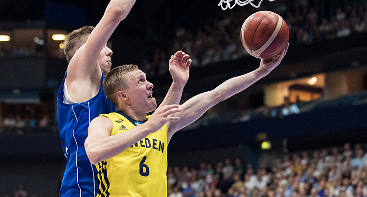 En kille i gult linne försöker göra ett skott i basket. En kille i blått linne försöker stoppa det.