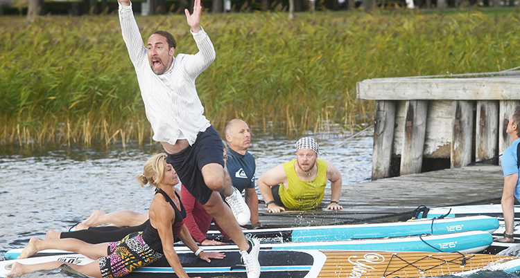 Han halkar ut i vattnet, sträcker händerna upp i luften. Bakom syns förvånade personer som ligger på surfingbrädor i vattnet.