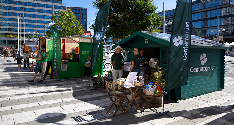 Flera små stugor står på ett torg. Det är olika politiska symboler på stugorna. Det står vimplar utanför stugorna. Folk är vid stugorna.