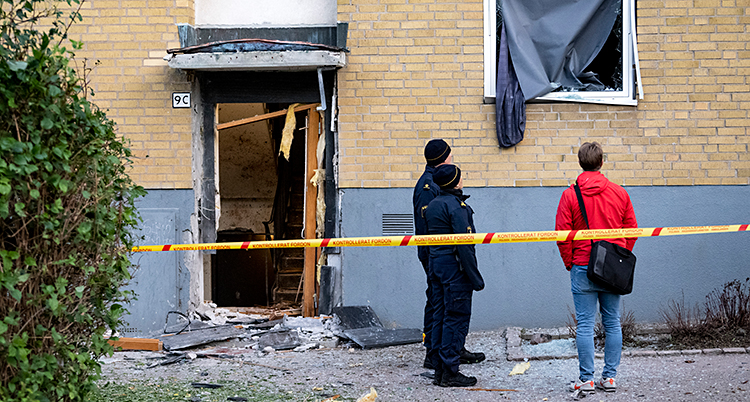 Två poliser och en tredje person står och tittar på ett lägenhetshus. Dörren och fönstret har blivit skadat.