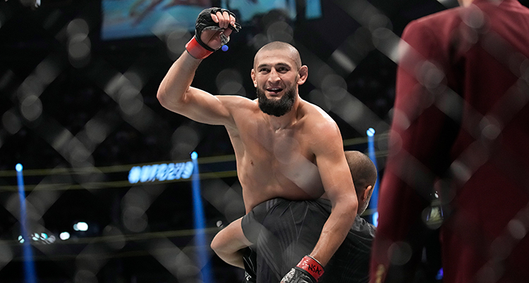 Bilden i tagen i en ring för kampsport. En man lyfter upp Khamzat Chimaev i luften. Khamzat är glad och sträcker armen i luften.