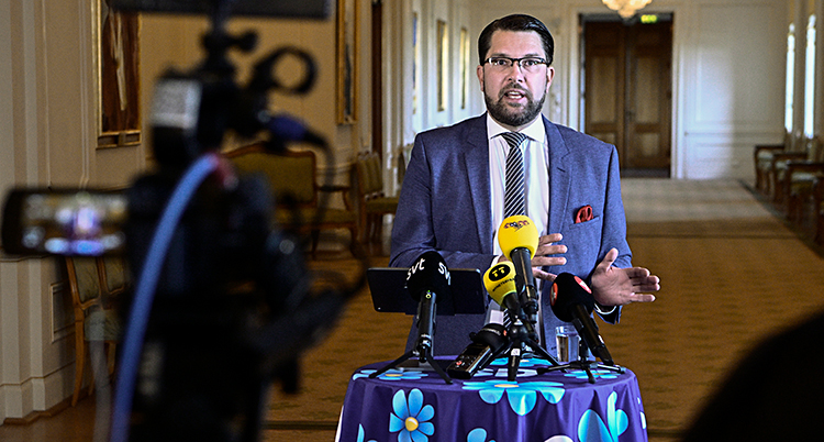 Jimmie Åkesson står vid ett bord och pratar. På bordet står flera mikrofoner.