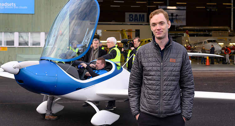 Han står längst fram. Bakom syns ett flygplan med öppen cockpit. Där sitter en man. Flera personer står runt flygplanet.