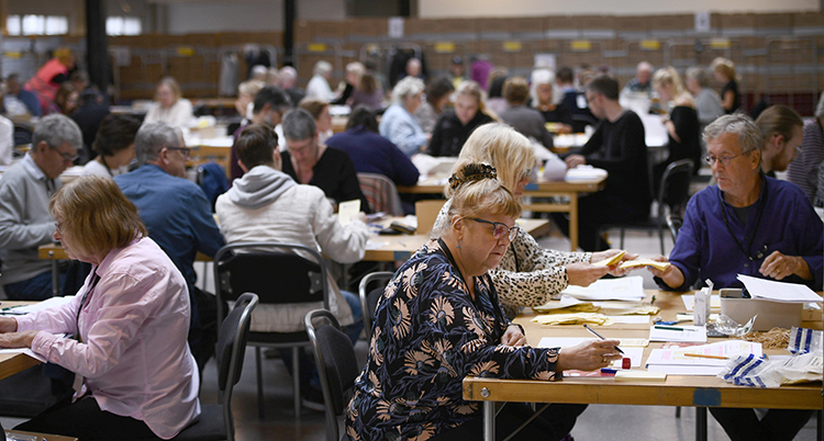 Människor vid långa bord sitter och räknar med massor av valsedlar framför sig.