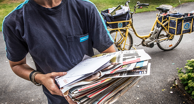 En brevbärare hålla ri en stor hög med reklam. Bakom står en postcykel.