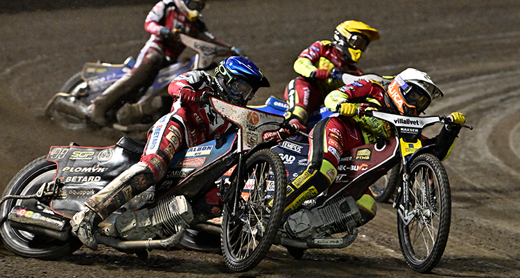Fyra speedwaymotorcyklar kör på en speedwaybana. Förarna har hjälmar och särskilda kläder. De lutar motorcyklarna.